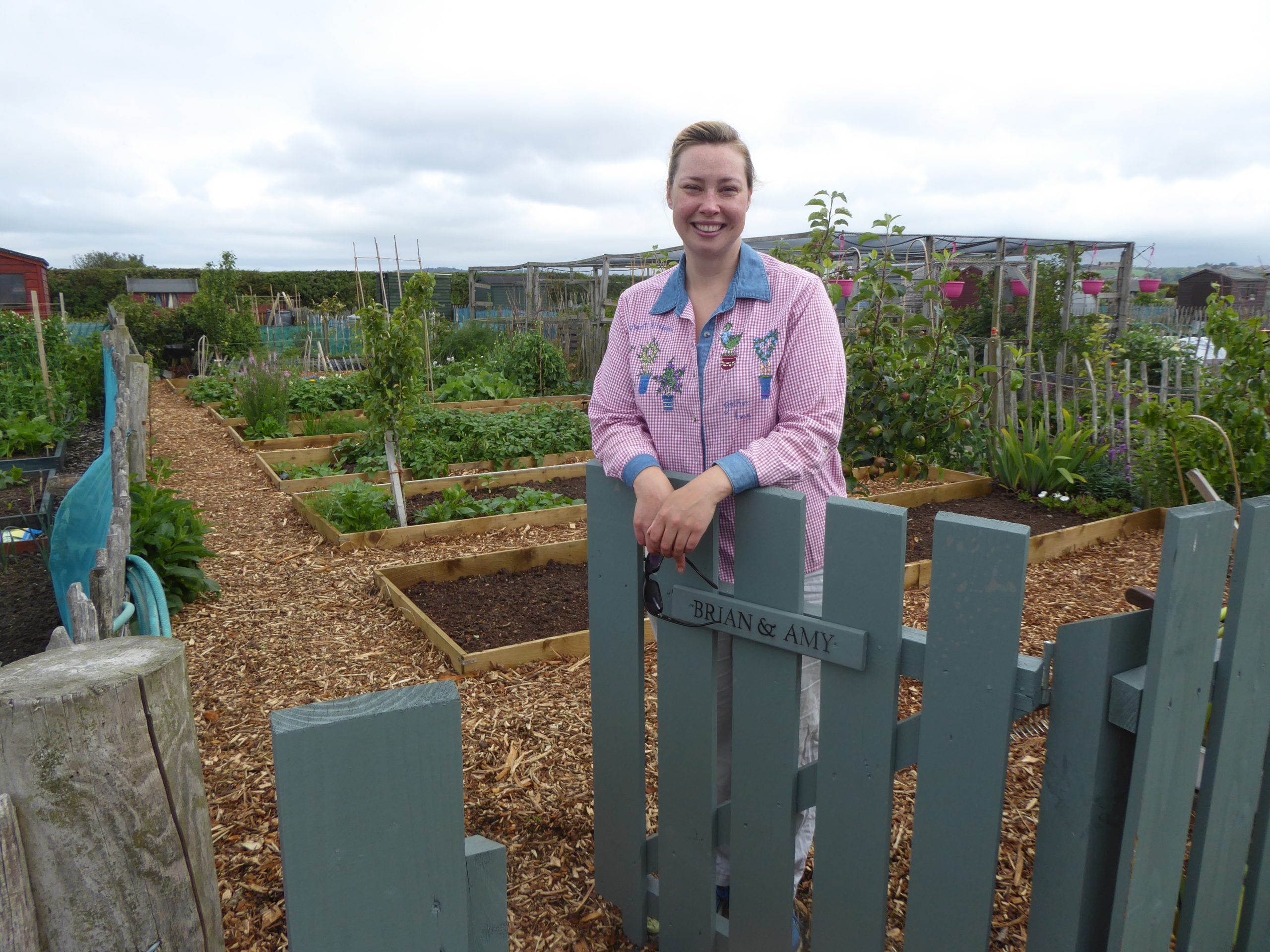 Amy Kelly standing at her allotment fence with vegetable and fruit beds behind her